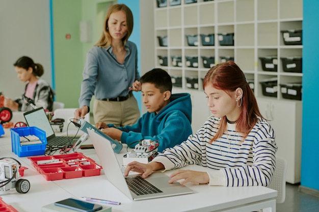 A middle school classroom scene where students are working collaboratively on a robotics project. They are using laptops to program their robots, and their facial expressions show concentration and teamwork. The teacher is observing and providing guidance.