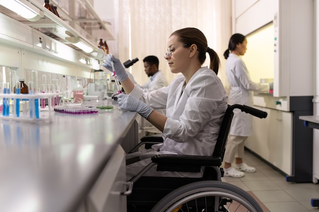 A student using a wheelchair is participating in a science class. The lab is equipped with adjustable workstations and accessible equipment. The student is working alongside their peers, conducting an experiment with the support of a teacher. The scene emphasizes inclusivity and equal access to STEM education.