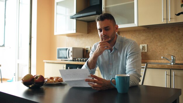 A man reviewing his financial documents at his kitchen table. The documents include expense reports and an insurance policy summary, with a calculator nearby.