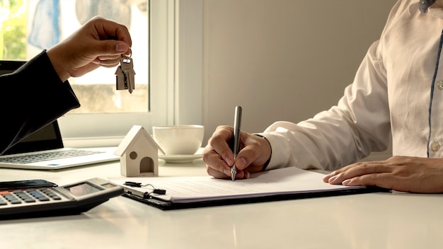 A close-up of a hand signing a mortgage document, with a pen and a calculator visible on the table. The background is blurred, suggesting a busy office environment.