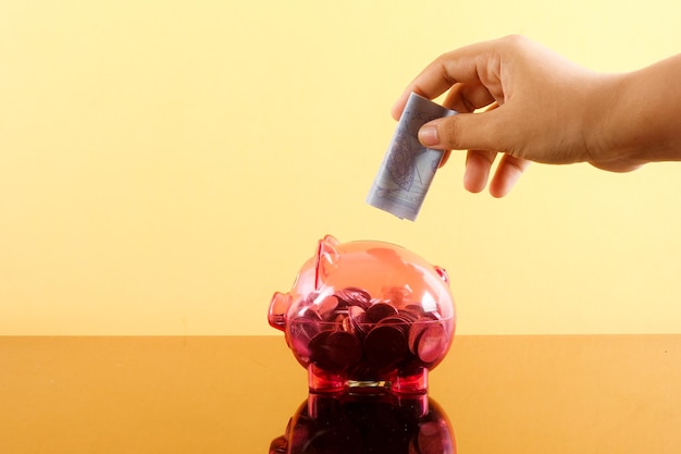 A close-up of hands carefully placing dollar bills into a piggy bank, set against a backdrop of financial charts and graphs. The image emphasizes the importance of consistent savings habits in retirement planning and maximizing 401(k) contributions.