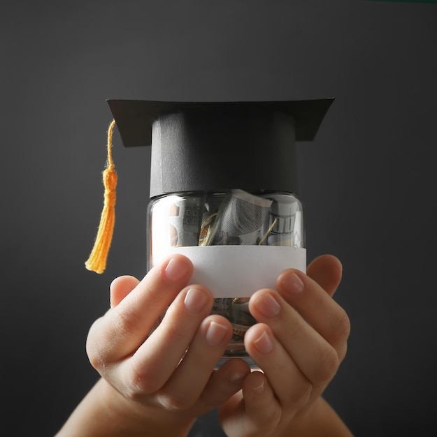 A person holding a stack of money with a blurred graduation cap in the background. The money is brightly lit.