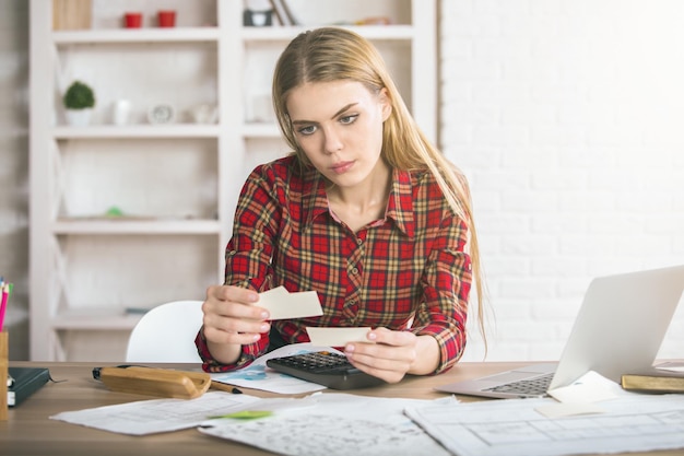 A woman sitting at a desk, using a calculator and reviewing financial documents. She appears focused and determined, with a laptop and notepad nearby.