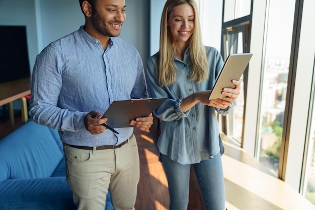 Two professionals in a brightly lit office, engaged in a friendly discussion, reviewing a tablet with graphs and charts displayed. One is pointing to the screen, emphasizing a point. Smiling, positive interaction, teamwork environment.